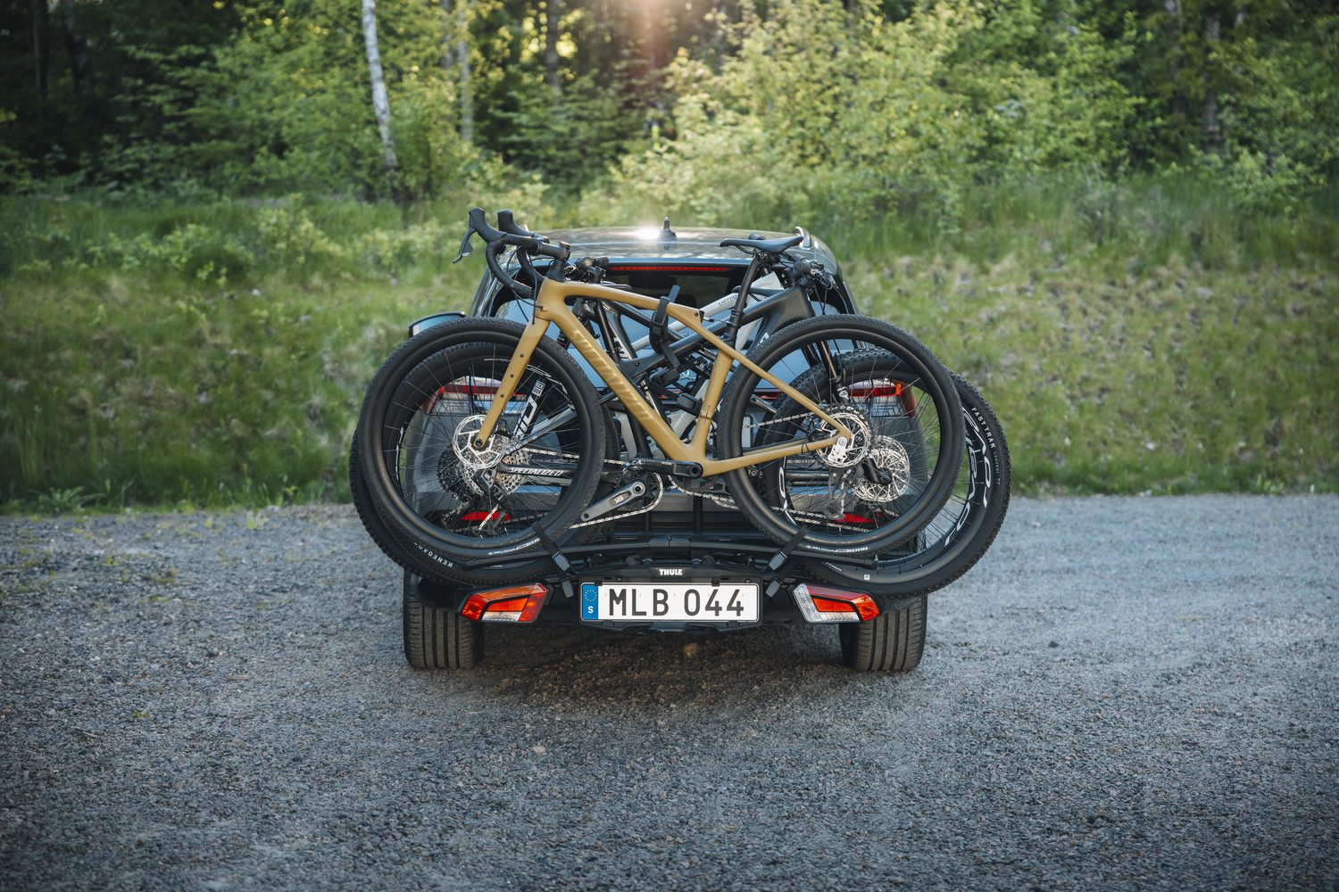 Drie fietsen staan op een Thule EasyFold 3 fietsendrager die achterop een auto is bevestigd die geparkeerd staat voor een boslandschap.