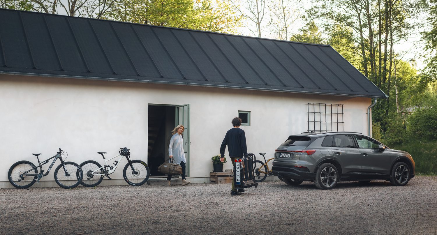 Een man in vrijetijdskleding draagt een fietsenrek aan het handvat en loopt tussen een huis en een SUV die geparkeerd staat op een oprit met grind.