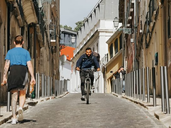 Twee personen, waarvan eentje fietsend en de andere lopend, in een straat in Lissabon.